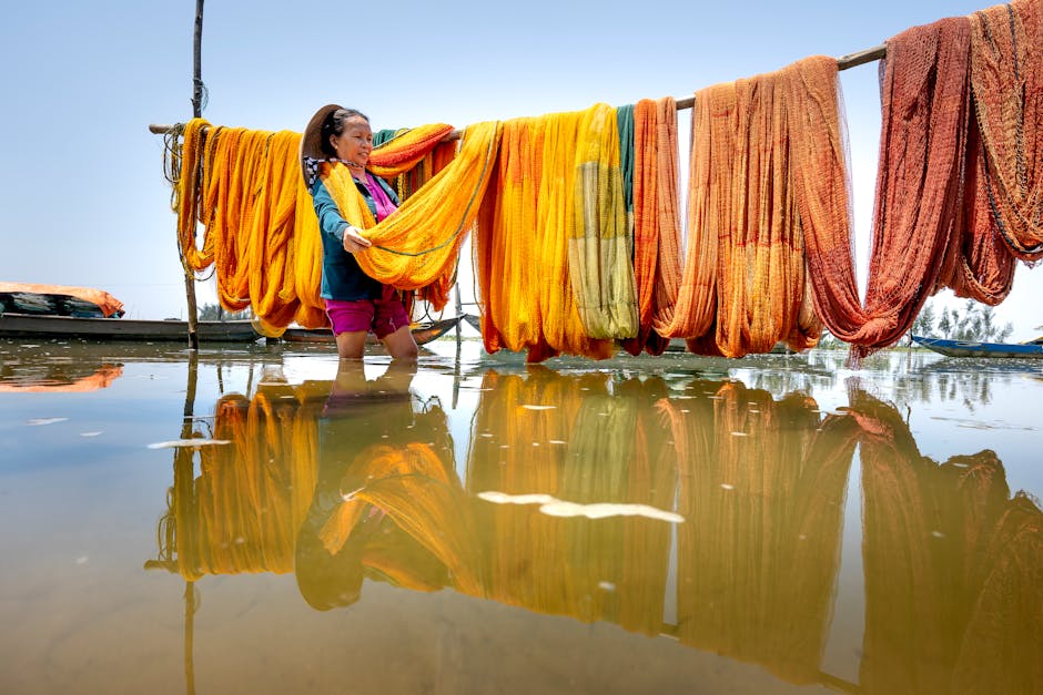 Positive middle aged Asian female in casual clothes standing in river water and choosing colorful fishing net hanging on bamboo pole on sunny day in Thua Thien Hue Province