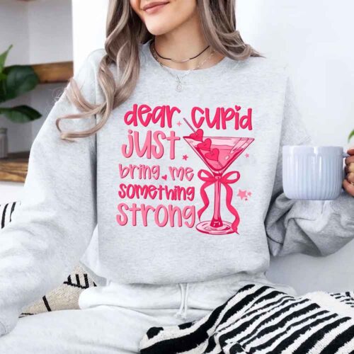 Woman in a gray sweatshirt with pink text and a martini graphic, holding a mug.